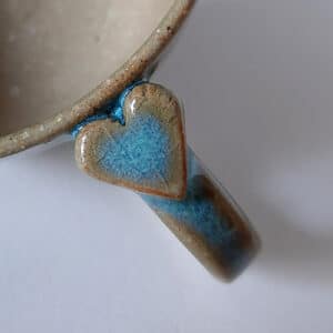 Détail. Petit déjeuner amoureux. En grès clair, émaillé de transparent, cette tasse est bordée d'un liseré couleur terre accompagné d'un bandeau dans les tons bleus céleste. Sa hanse vous offre un joli cœur à sa base.