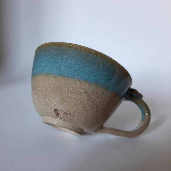 De face. Petit déjeuner amoureux. En grès clair, émaillé de transparent, cette tasse est bordée d'un liseré couleur terre accompagné d'un bandeau dans les tons bleus céleste. Sa hanse vous offre un joli cœur à sa base.