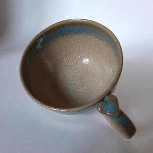 Le fond. Petit déjeuner amoureux. En grès clair, émaillé de transparent, cette tasse est bordée d'un liseré couleur terre accompagné d'un bandeau dans les tons bleus céleste. Sa hanse vous offre un joli cœur à sa base.