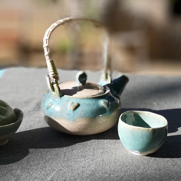 Théière en grés émaillé avec anse en osier et ruban synthétique. Collection Lagon, un émail transparent accompagné d'un nuage turquoise sur le bord de la pièce. La transparence de l'émail apporte une légèreté. La couronne de cœur apporte une touche de romantisme. Elle possède un filtre incorporé ce qui permet de filtrer toute sorte de thé.
