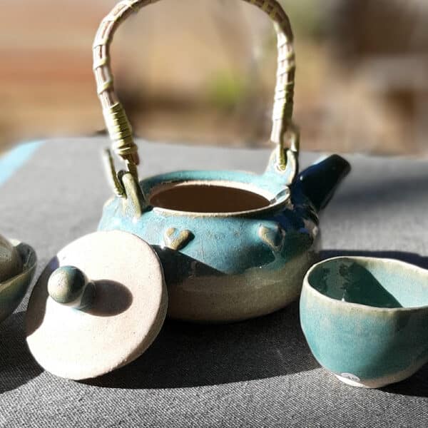 Théière en grés émaillé avec anse en osier et ruban synthétique. Collection Lagon, un émail transparent accompagné d'un nuage turquoise sur le bord de la pièce. La transparence de l'émail apporte une légèreté. La couronne de cœur apporte une touche de romantisme. Elle possède un filtre incorporé ce qui permet de filtrer toute sorte de thé.