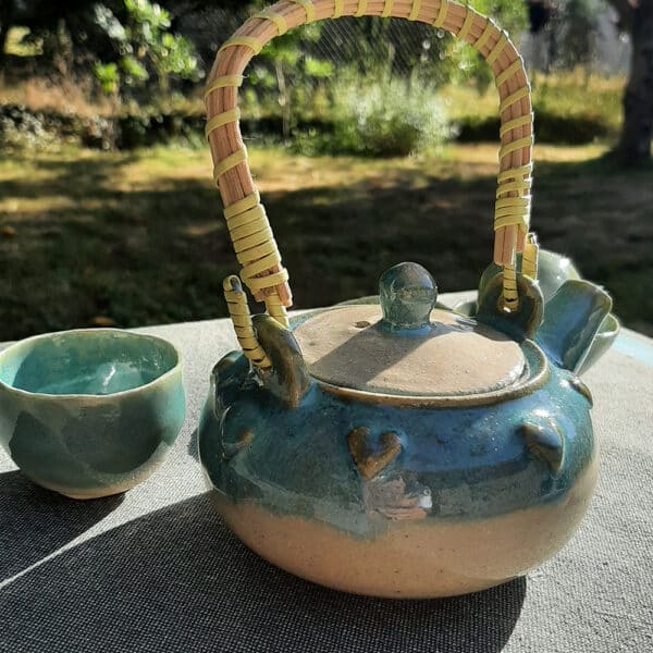 Théière en grés émaillé avec anse en osier et ruban synthétique. Collection Lagon, un émail transparent qui diffuse un ruban turquoise sur le bord de la pièce. La transparence de l'émail apporte une légèreté. La couronne de cœur apporte une touche de romantisme. Elle possède un filtre incorporé ce qui permet de filtrer toute sorte de thé.