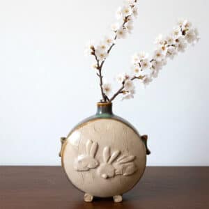 Dans la collection des "Elégantes", vase en grés émaillé Azurite avec motif de lapin sur face avant sur fond transparent. Céramique aux 2 couleurs bleu et marron sur l'arrière, au dos et côté avec effet de matière. Mise en situation avec bouquet Ikebana (branche de cerisier en fleur).