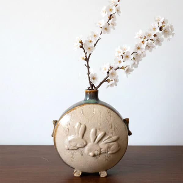 Dans la collection des "Elégantes", vase en grés émaillé Azurite avec motif de lapin sur face avant sur fond transparent. Céramique aux 2 couleurs bleu et marron sur l'arrière, au dos et côté avec effet de matière. Mise en situation avec bouquet Ikebana (branche de cerisier en fleur).