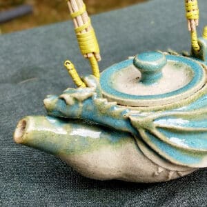 Détail du dragon. Théière en grés émaillé avec anse en osier et ruban synthétique. Collection Lagon, un émail transparent accompagné d'un nuage turquoise sur le bord de la pièce. La transparence de l'émail apporte une légèreté. Le turquoise vient souligner la forme du dragon. Elle possède un filtre incorporé ce qui permet de filtrer toute sorte de thé.